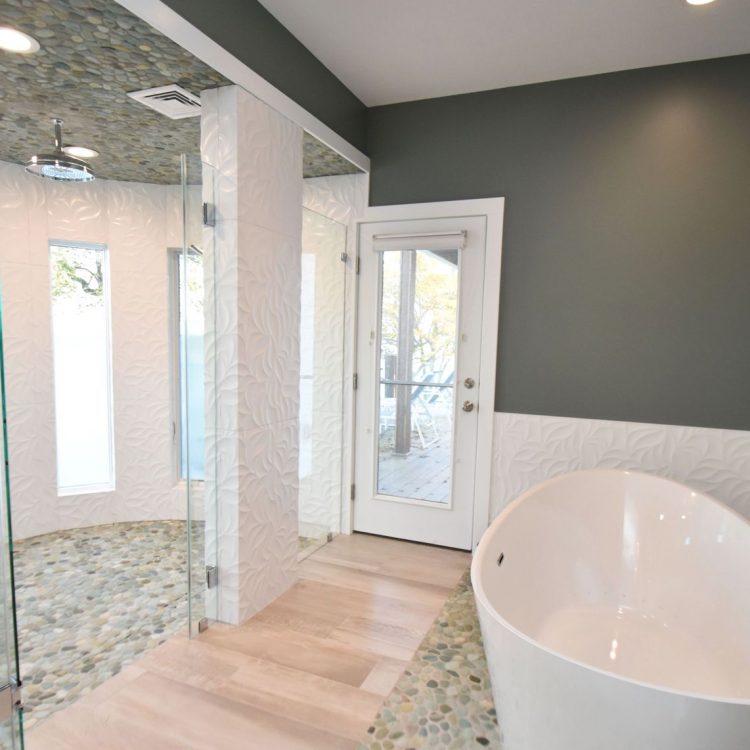 bath remodeling rochester bath and kitchen - A large circular shower with slender rectangular frosted window panels. The ground has a blue green pebble mosaic and the walls are white with leaf shaped texture to the right of the shower is a large standalone tub